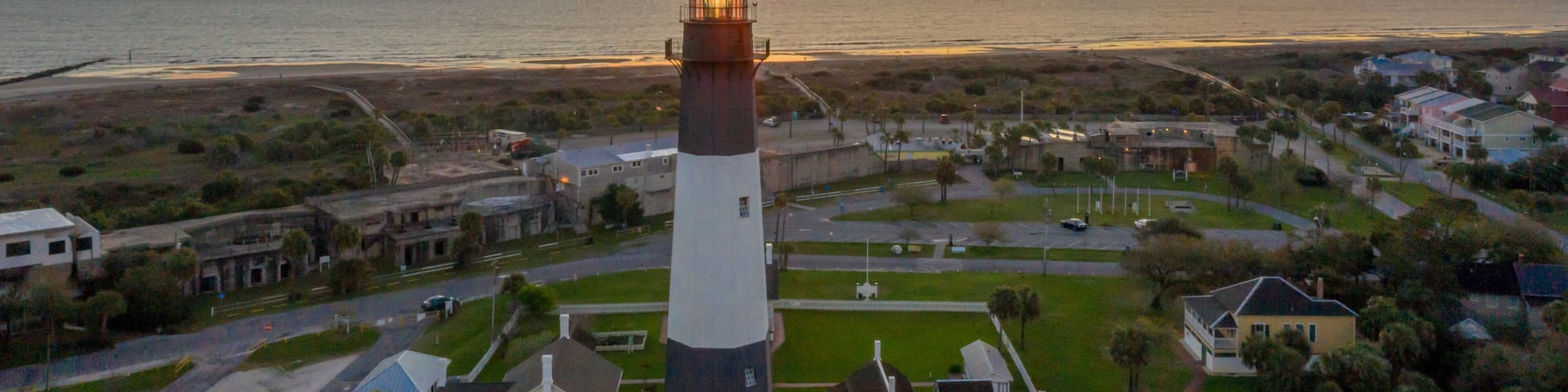 Un pueblo costero al atardecer, con un faro destacado, playa y zona residencial.