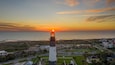 A coastal town at sunset with a prominent lighthouse, beach, and residential area.