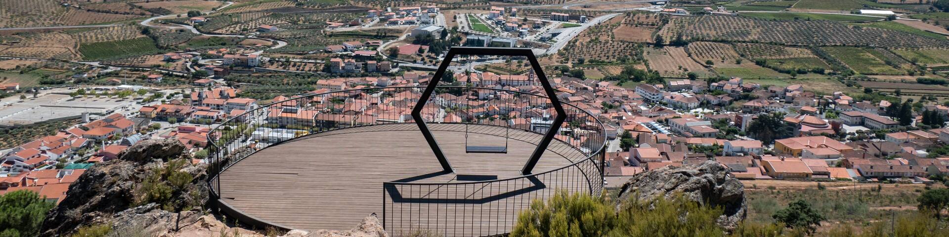 Miradouro redondo com estrutura metálica e com o chão em madeira e com um baloiço em formato hexagonal junto às capelinhas em Vila Flor e com a vila ao fundo em Portugal