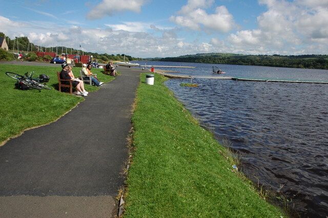 A riverside path with grass on one side and water on the other, bicycles, and people sitting and walking.