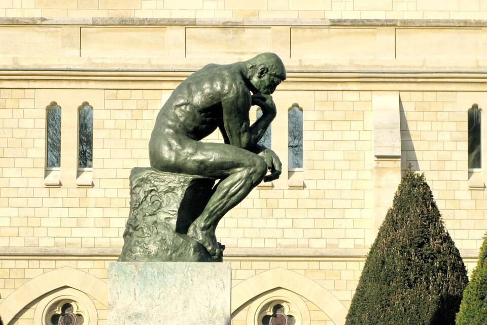 A bronze statue of a contemplative man on a pedestal in front of a building with arched windows and a neatly trimmed hedge.