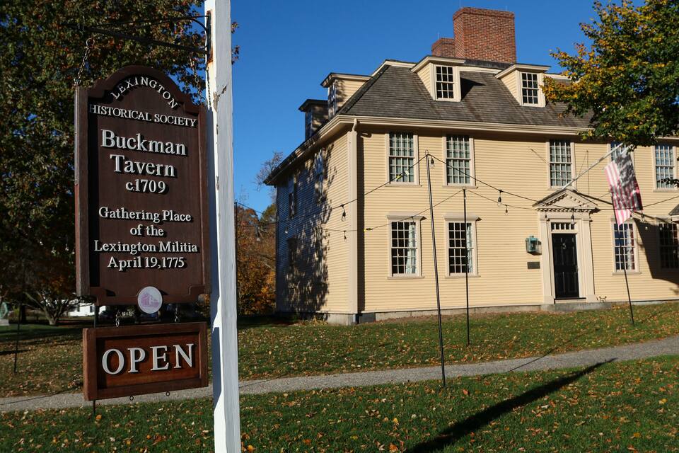 A historical building with a sign reading "Buckman Tavern c.1709 Gathering Place of the Lexington Militia April 19, 1775" and an "OPEN" sign below it.