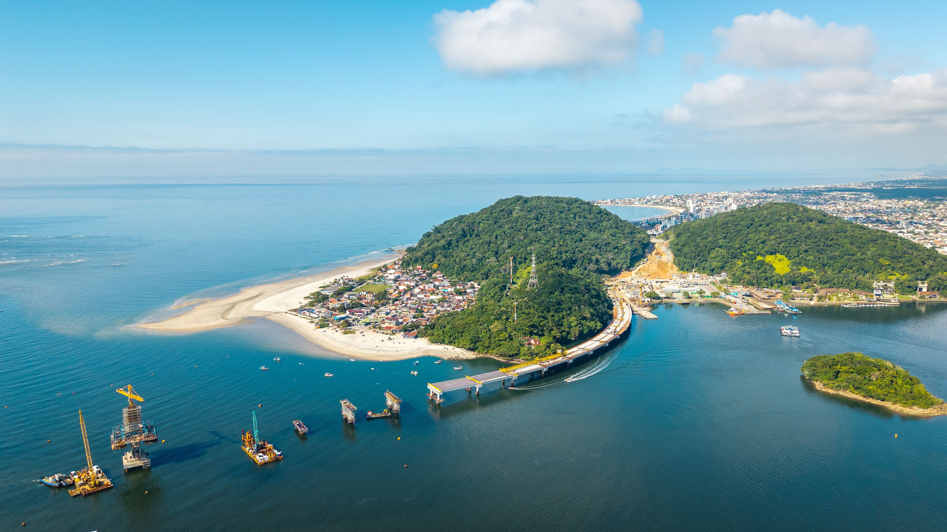 Wide aerial view of Guaratuba-Matinhos bridge construction with sandy beaches, forested hills and Guaratuba city in the background.