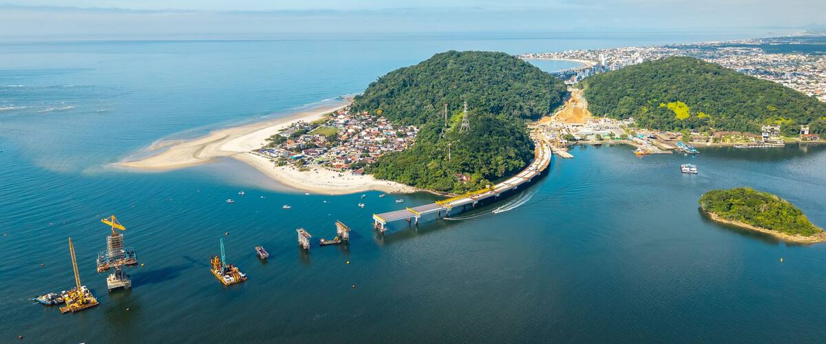 Wide aerial view of Guaratuba-Matinhos bridge construction with sandy beaches, forested hills and Guaratuba city in the background.