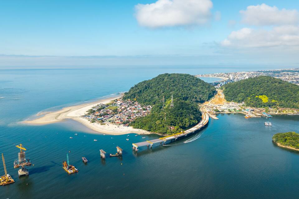 An aerial view of a coastal area with a bridge connecting two landmasses, a beach, and a forested hill.