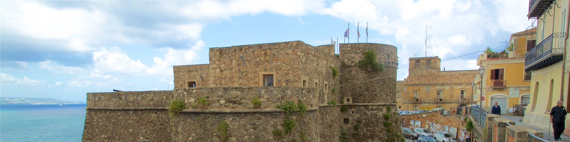 A stone fortification overlooking a coastal road with parked cars and a stop sign.
