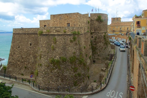 Une fortification en pierre surplombant une route côtière, où sont garées des voitures et où se trouve un panneau de signalisation.