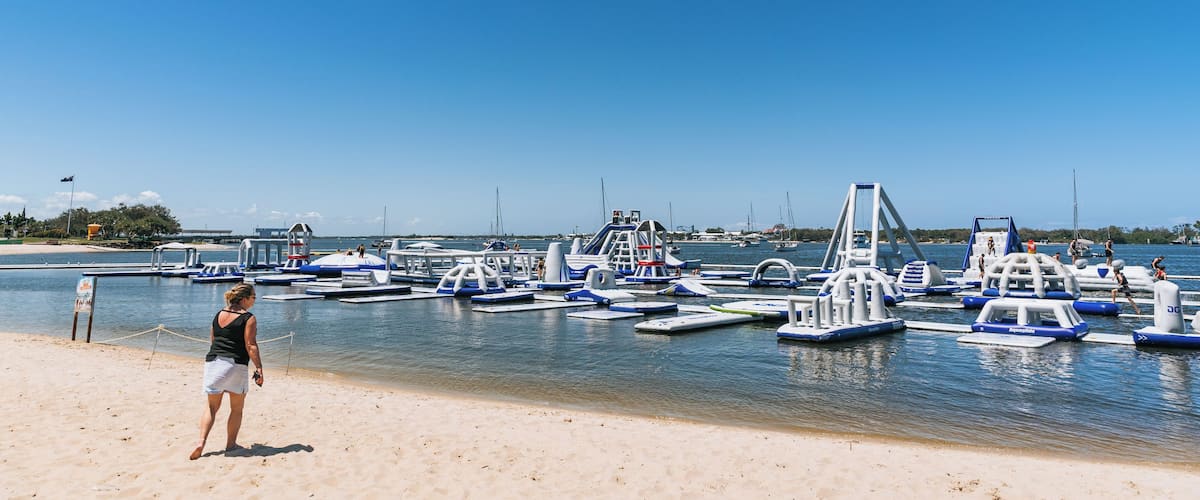 Une plage avec un ciel dégagé, une femme debout sur le sable, et plusieurs embarcations bleues et blanches amarrées dans l’eau.
