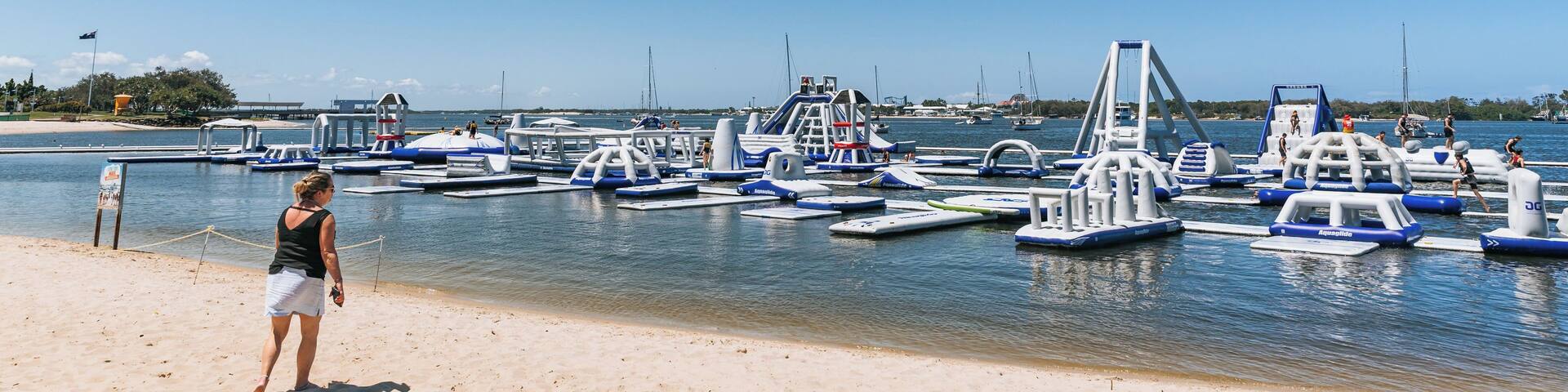 A beach with a clear sky, a woman standing on the sand, and several blue and white watercraft docked in the water.