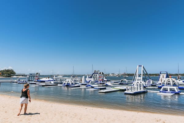 Ein Strand mit blauem Himmel, eine Frau, die am Sand steht, und mehrere blau-weiße Wasserfahrzeuge, die im Wasser liegen.