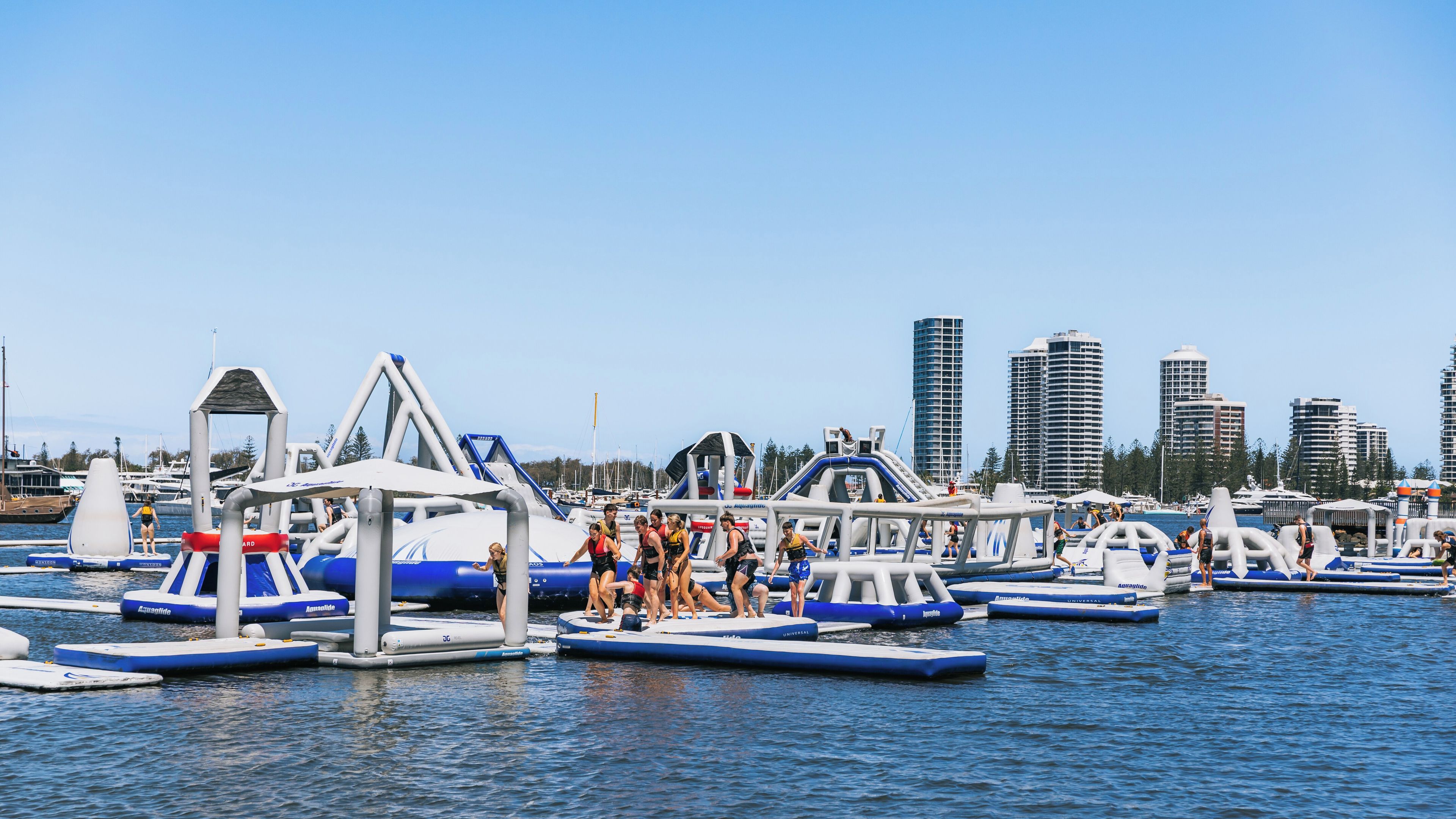 An inflatable water park with various play structures on the water, surrounded by a marina with boats and tall buildings in the background.