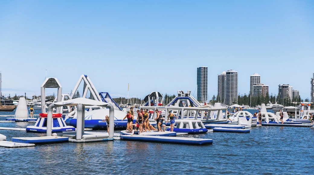 An inflatable water park with various play structures on the water, surrounded by a marina with boats and tall buildings in the background.