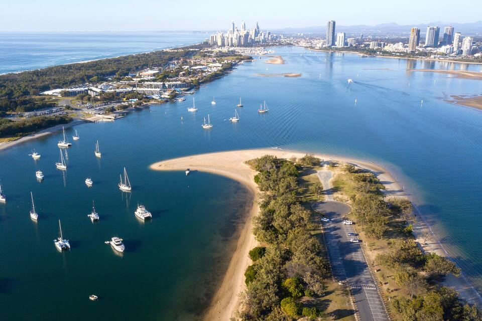 Vista aérea de una ciudad costera con un puerto deportivo lleno de veleros, una playa arenosa y una zona urbana densa con rascacielos al fondo.