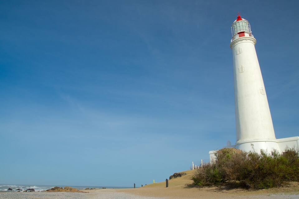 A tall white lighthouse with a red top, situated on a sandy beach with rocky outcrops and sparse vegetation.