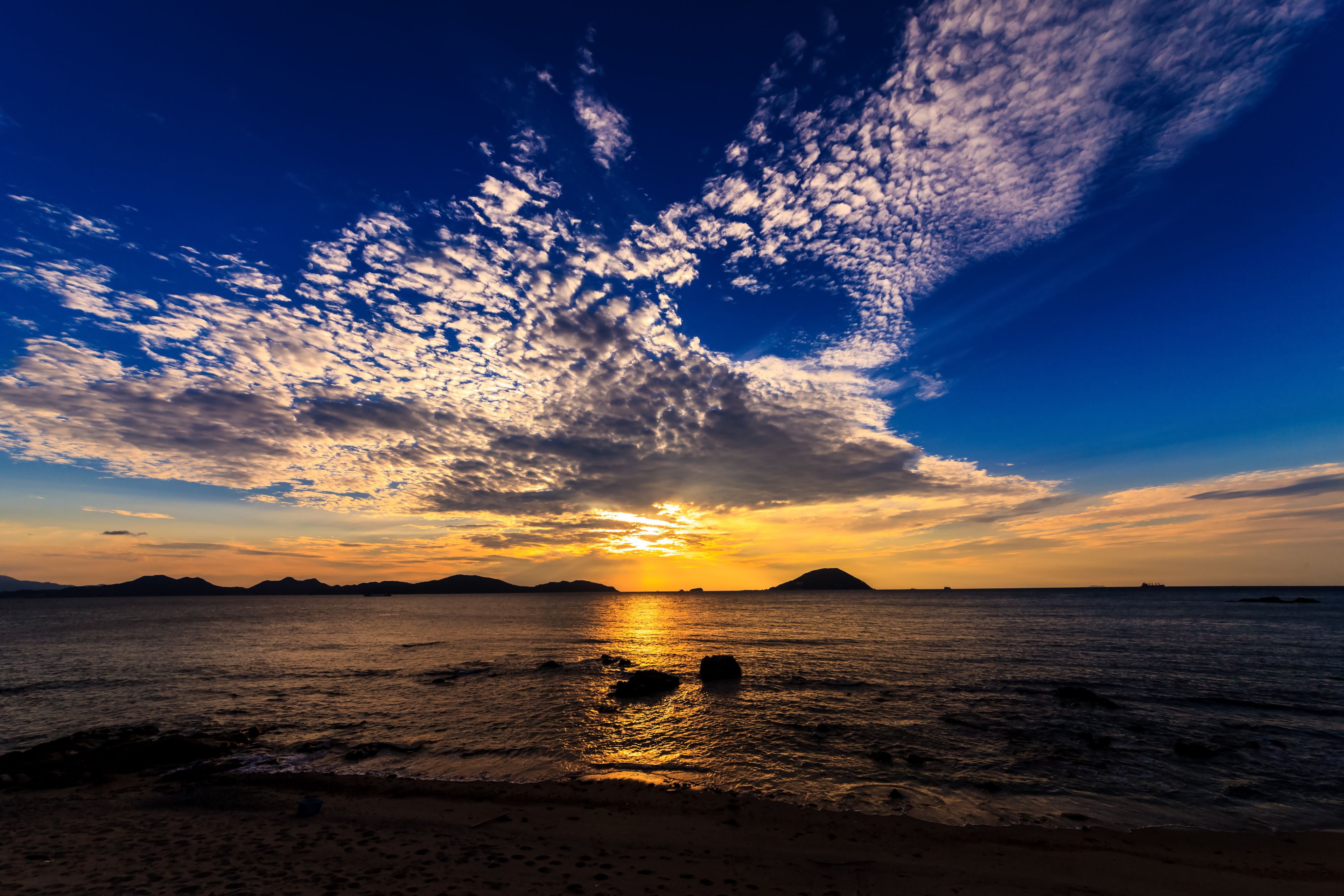A sunset over a beach with waves, a distant island, and scattered clouds.