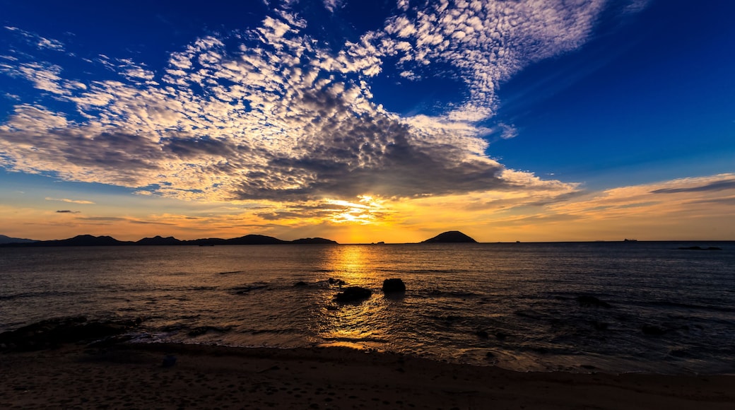 A sunset over a beach with waves, a distant island, and scattered clouds.