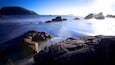 A rocky coastline with mist over the water and distant hills.