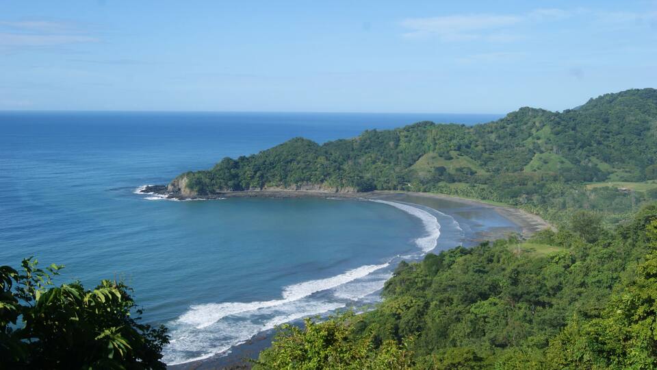 A coastal landscape with a curved bay, dense green forest, and a clear blue ocean.