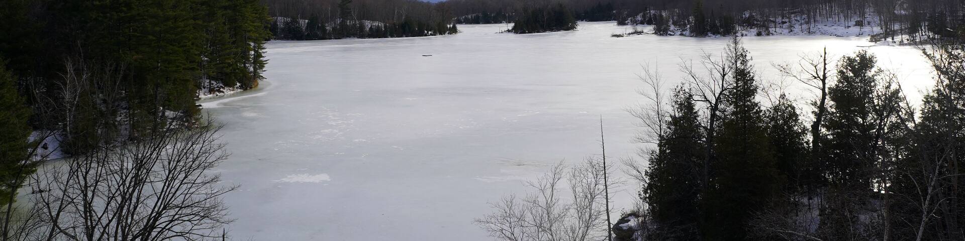 A wide, frozen lake is surrounded by dense evergreen and deciduous forests under a vast, cloud-filled sky near Sharbot Lake, Ontario