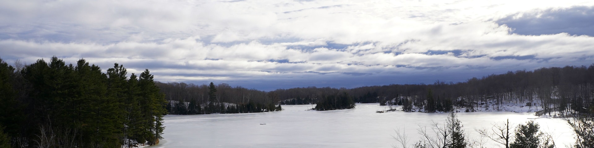 A wide, frozen lake is surrounded by dense evergreen and deciduous forests under a vast, cloud-filled sky near Sharbot Lake, Ontario
