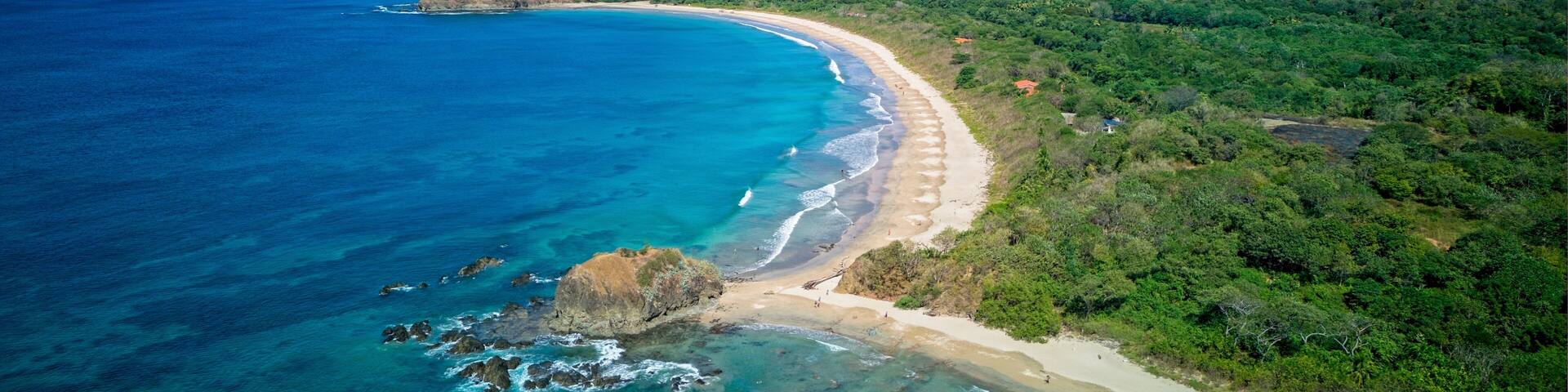 A coastal landscape with a sandy beach, clear blue waters, and green hills in the background.