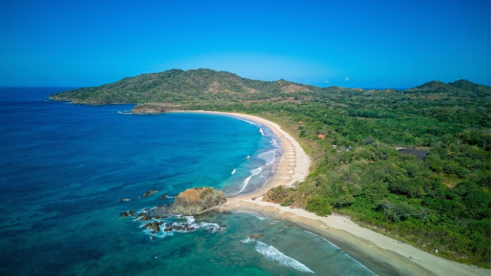 Un paysage côtier avec une plage de sable, des eaux d’un bleu limpide et des collines verdoyantes en arrière-plan.