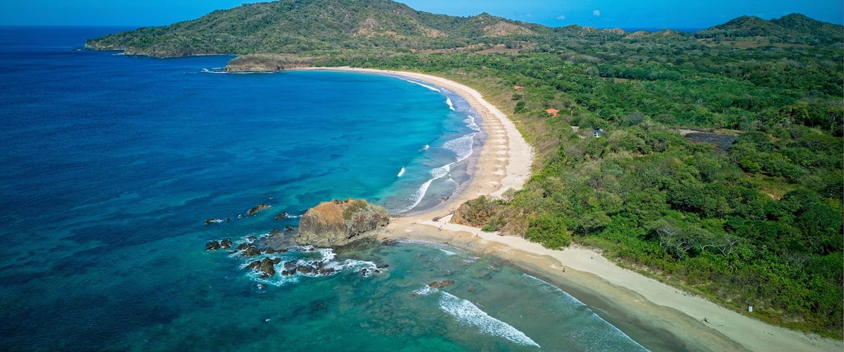 A coastal landscape with a sandy beach, clear blue waters, and green hills in the background.
