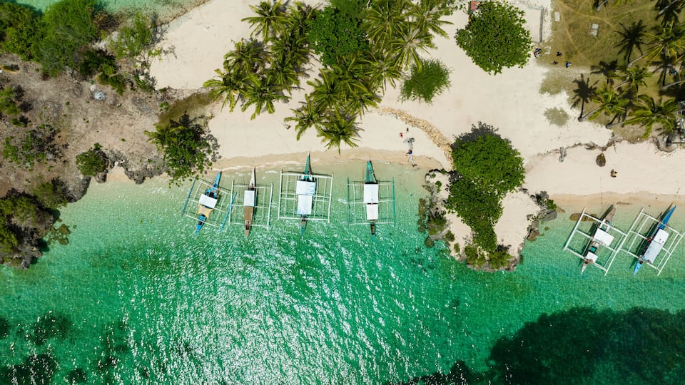An aerial view of a beach with clear turquoise water, palm trees, and several small boats anchored near the shore.