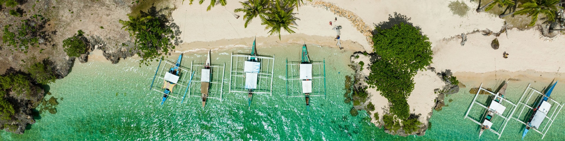 Vista aérea de una playa con agua turquesa cristalina, palmeras y varias embarcaciones pequeñas ancladas cerca de la orilla.