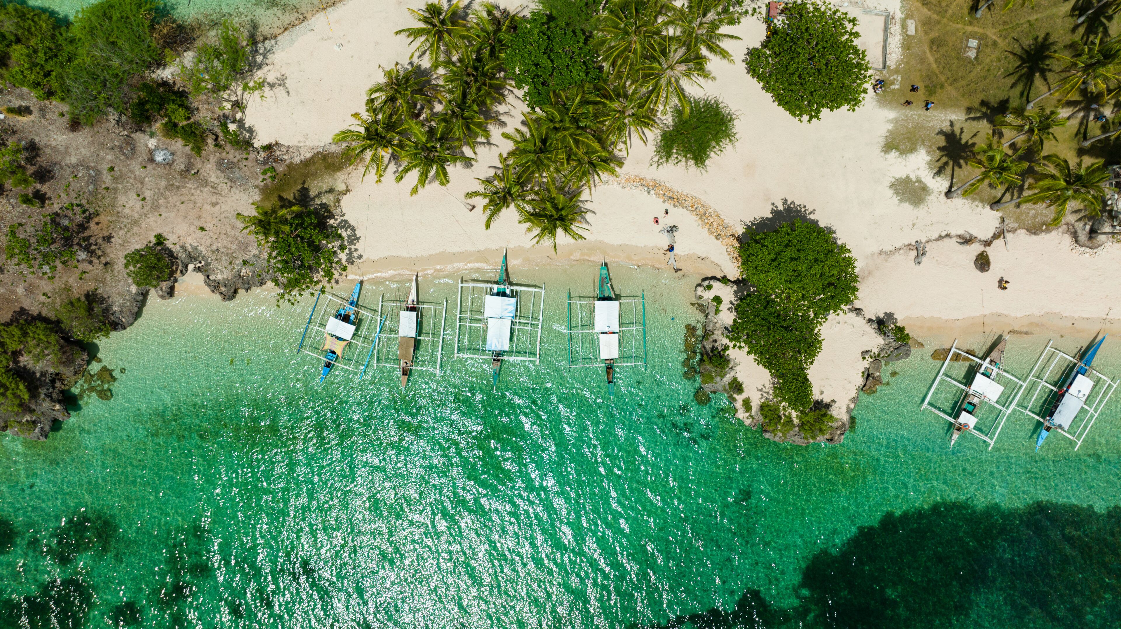 An aerial view of a beach with clear turquoise water, palm trees, and several small boats anchored near the shore.