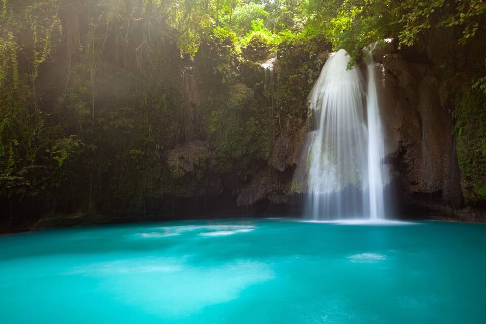 A turquoise pool with a waterfall cascading down a rocky cliff surrounded by lush greenery.