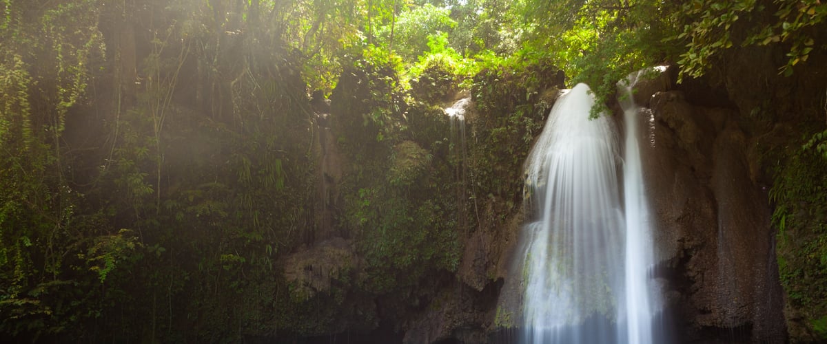 A turquoise pool with a waterfall cascading down a rocky cliff surrounded by lush greenery.