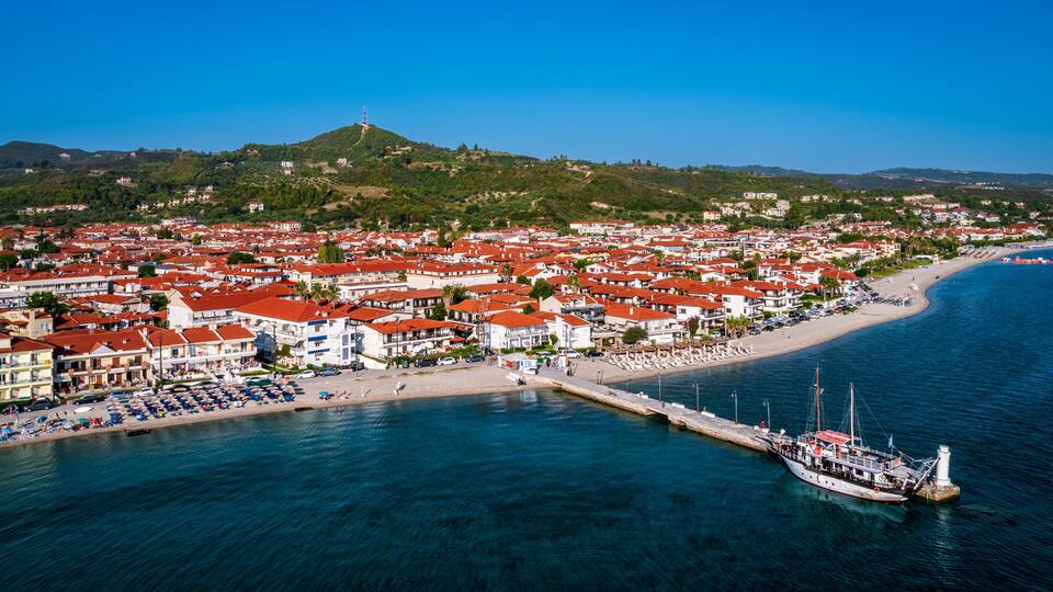 A coastal town with red-roofed buildings, a beach, and a lighthouse on a hill.