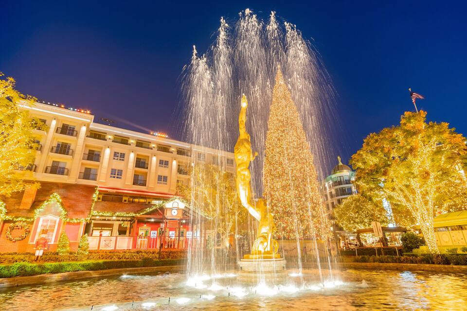 Uma fonte com jatos de água, uma estátua dourada e um pinheiro de Natal com luzes.