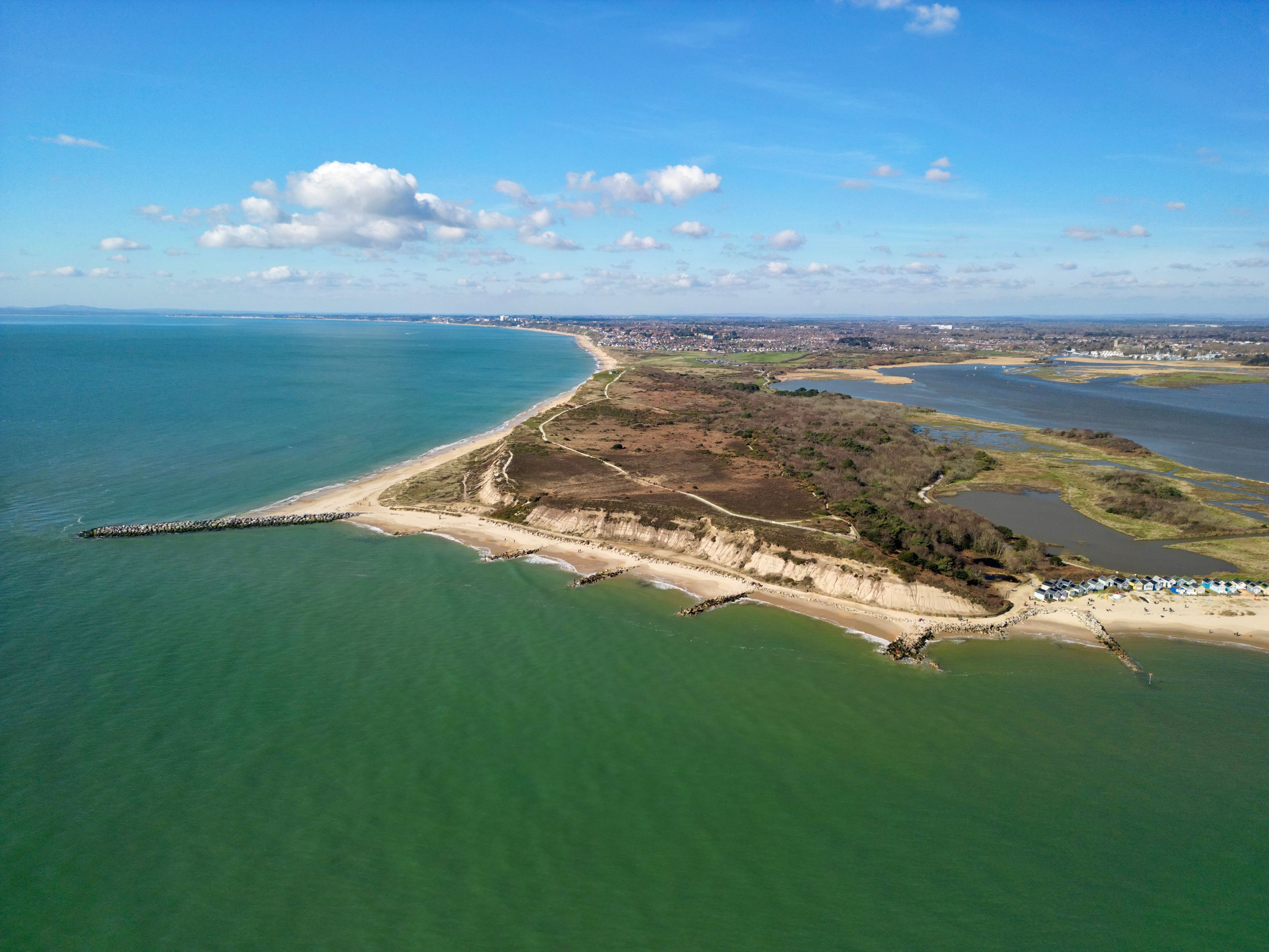 An aerial view of a coastal area with a sandy beach, a rocky breakwater, and a small settlement with buildings.