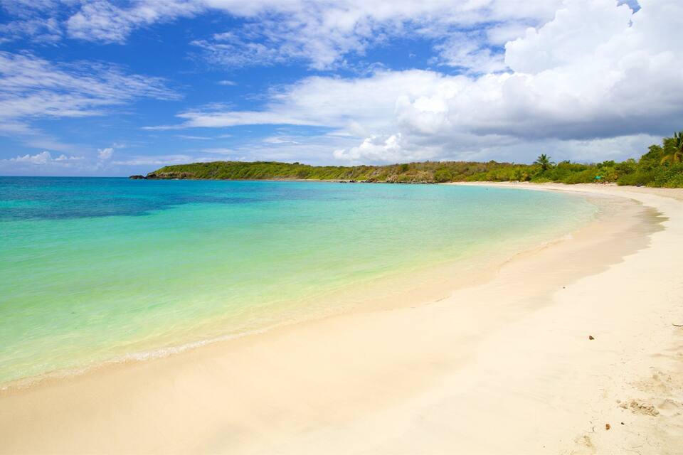 Uma praia com águas turquesa cristalinas, um areal dourado e colinas verdes ao fundo sob um céu parcialmente nublado.