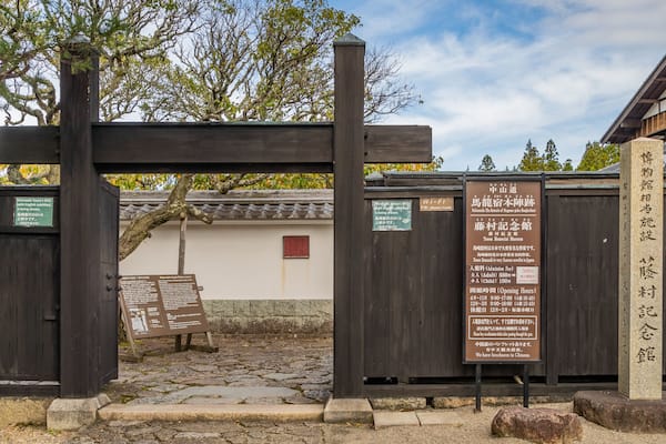 Une porte d’entrée japonaise traditionnelle, avec des piliers en bois et un chemin de pierre menant à un bâtiment doté d’un toit en tuiles.
