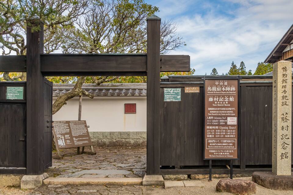 A traditional Japanese gate with wooden pillars and a stone pathway leading to a building with a tiled roof.