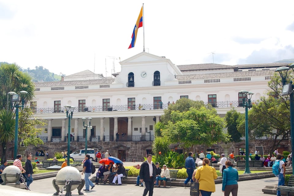 Un edificio blanco grande con una bandera, rodeado de gente y follaje.