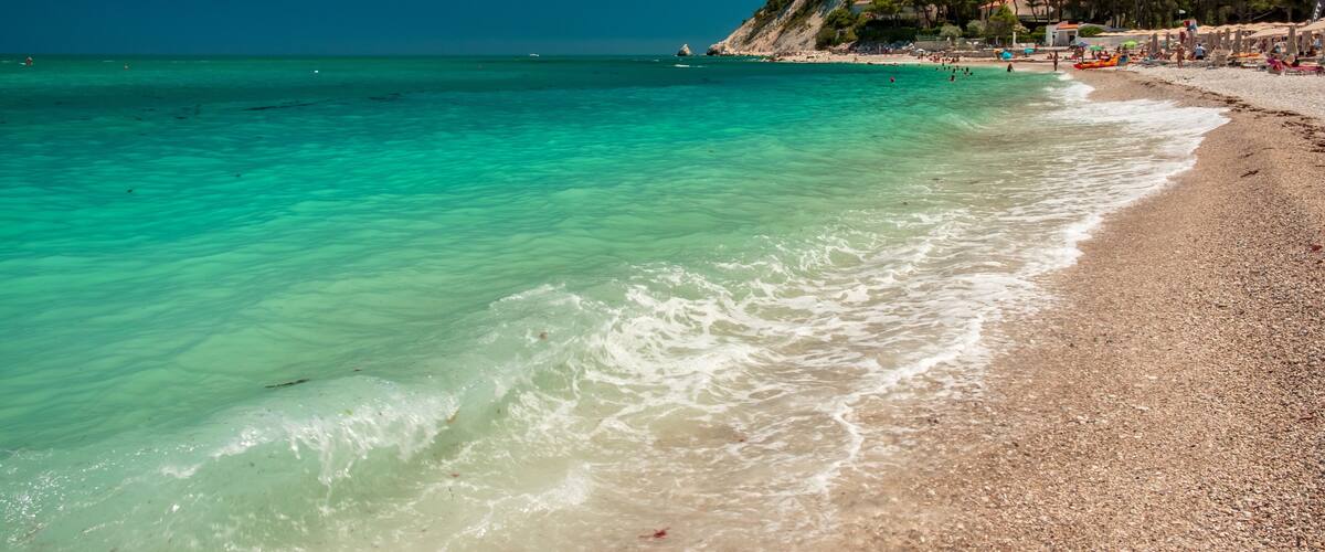 Une plage aux eaux turquoise, bordée de galets, et une colline parsemée de bùtiments.