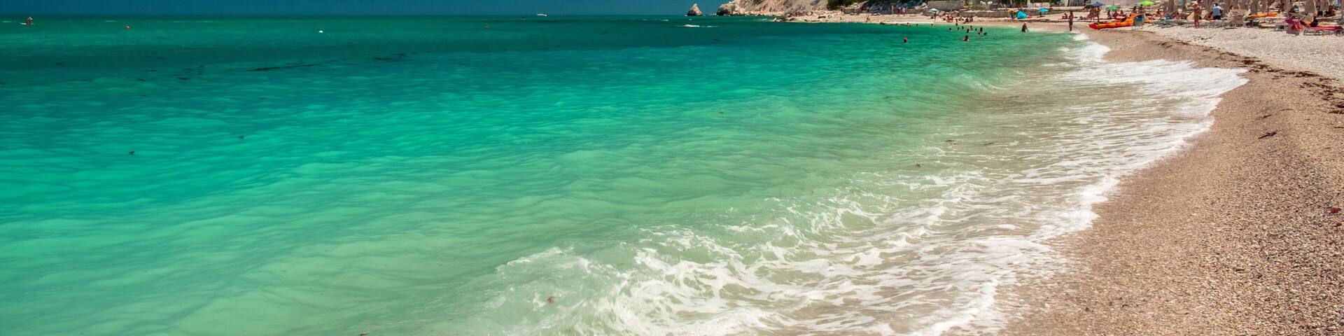 A turquoise beach with clear waters, pebbly shore, and a hillside with buildings.
