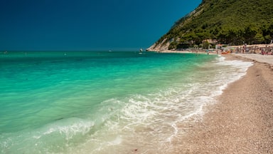 Ein türkisfarbener Strand mit klarem Wasser, kiesigem Ufer und einer Hügelkuppe mit Gebäuden.