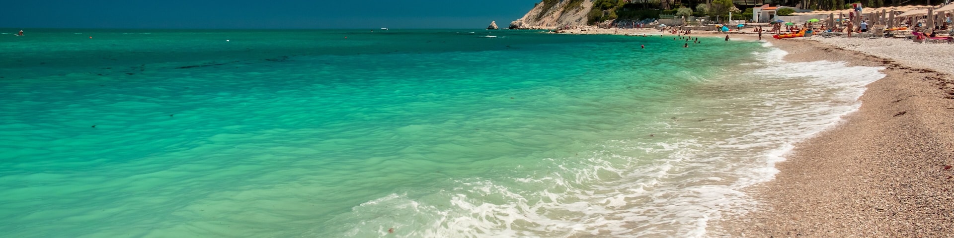 A turquoise beach with clear waters, pebbly shore, and a hillside with buildings.