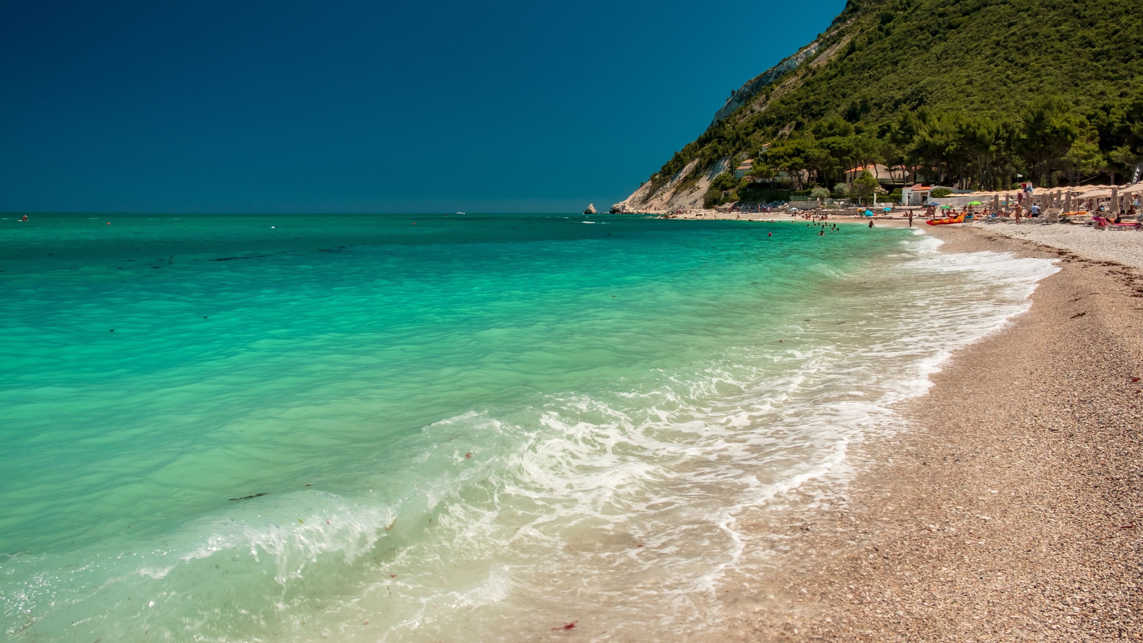 A turquoise beach with clear waters, pebbly shore, and a hillside with buildings.