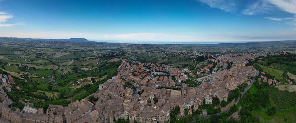 A panoramic view of a historic town with a central fortress, surrounded by green fields and distant mountains.