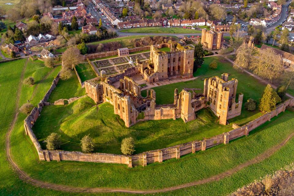An aerial view of a historic castle complex with surrounding green fields and a residential area in the background.