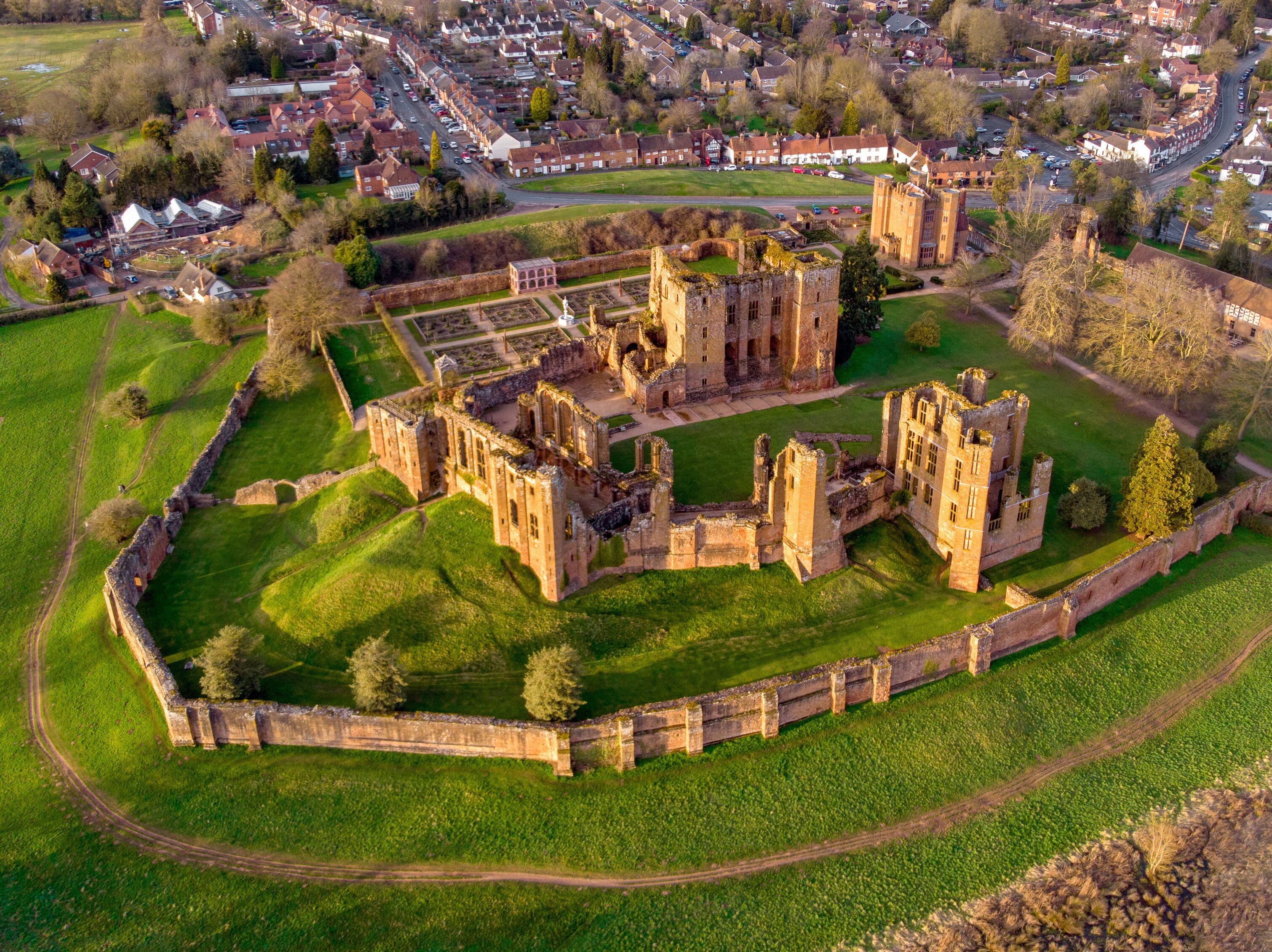 An aerial view of a historic castle complex with surrounding green fields and a residential area in the background.