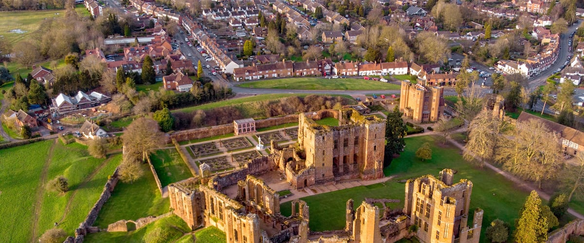 An aerial view of a historic castle complex with surrounding green fields and a residential area in the background.