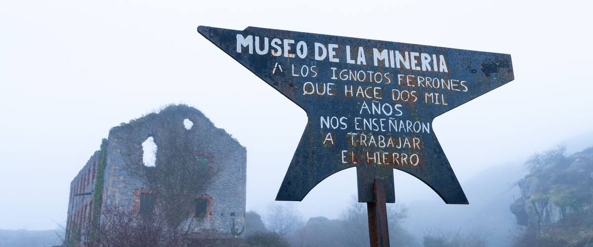 A signpost with text in Spanish, pointing towards a historical mining site with a dilapidated building in the background.