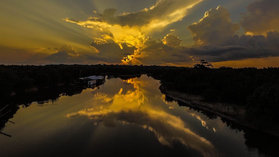A sunset over a calm river with a house on the left bank and dense trees in the background.